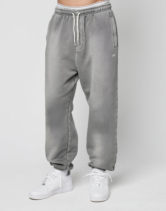 A person models the LIVE FAST DIE YOUNG Basic Loose Jogger—light gray, loose-fitting sweatpants with a logo print and white drawstrings—paired with white sneakers against a plain light gray background.
