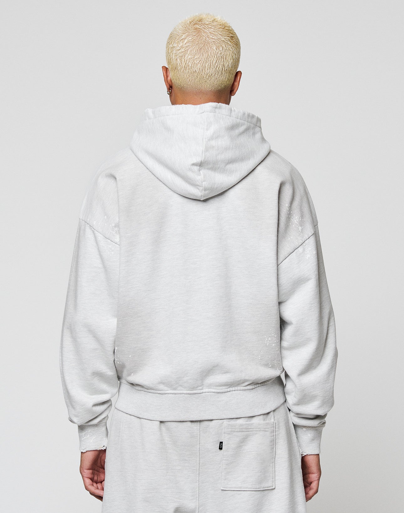 A person with short, bleached blonde hair wears the LIVE FAST DIE YOUNG Live Fast Rivet Hooded Zip in light gray, styled oversized, standing with their back to the camera against a plain light background.
