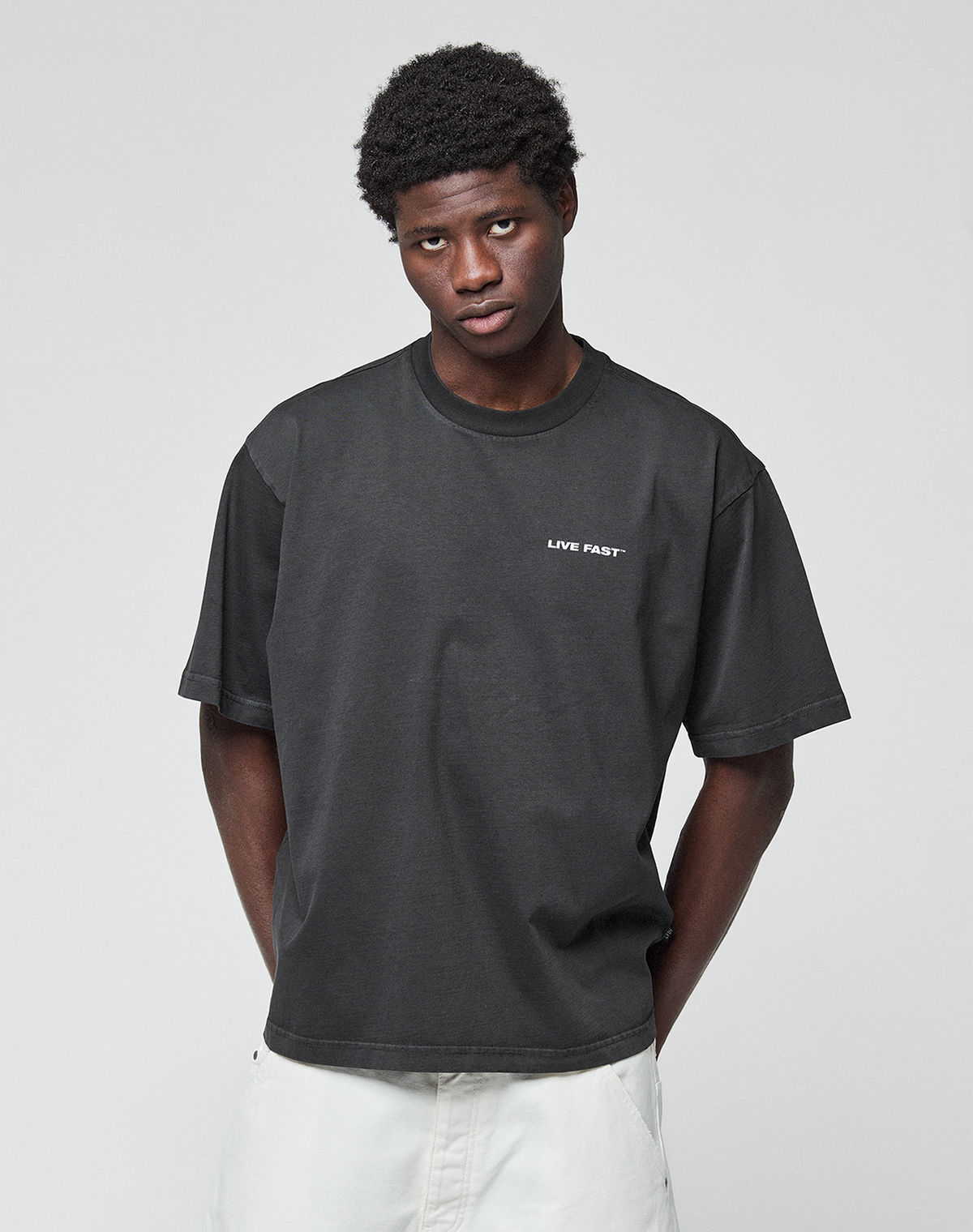 A man with short curly hair wears the LFDY Grand Theft Tee by LIVE FAST DIE YOUNG, a black 100% Baumwolle oversized T-shirt with LIVE FAST front and back print, paired with light-colored pants against a light gray background.