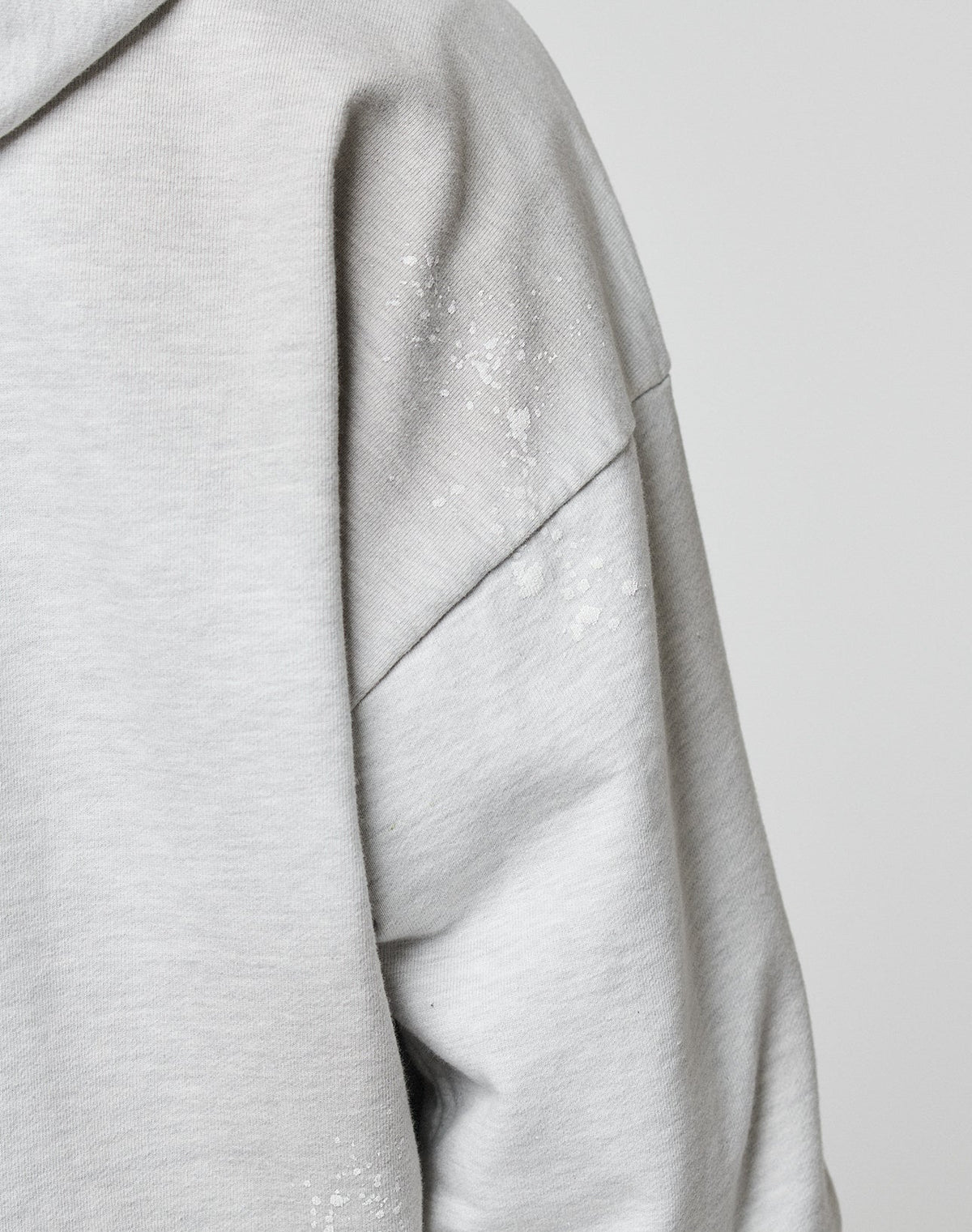 Close-up of the Live Fast Rivet Hooded Zip by LIVE FAST DIE YOUNG: light gray sleeve with handcrafted spray effect, small white paint specks on the shoulder, and modern distressing—all in a relaxed oversized fit against a plain background.