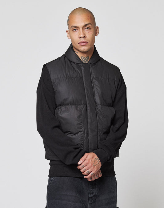 A person with a shaved head and visible tattoos wears a black LIVE FAST DIE YOUNG Basic Cropped Vest over a black sweater, standing against a plain light grey background with a neutral expression.