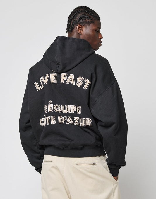 A person with braided hair wears the LIVE FAST DIE YOUNG LFDY LÉquipe Hooded Zip in black, featuring bold “LIVE FAST” and “LÉQUIPE CÔTE DAZUR” text on the back, paired with light pants against a plain background.