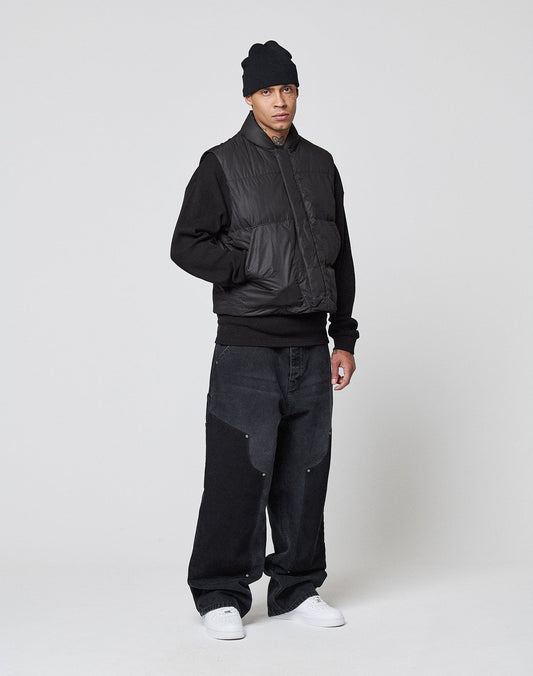 A man stands against a white background wearing a LIVE FAST DIE YOUNG Basic Cropped Vest over a Regular Fit black sweatshirt, baggy black jeans with reinforced knees and large pockets, a black beanie, and white sneakers, looking at the camera neutrally.