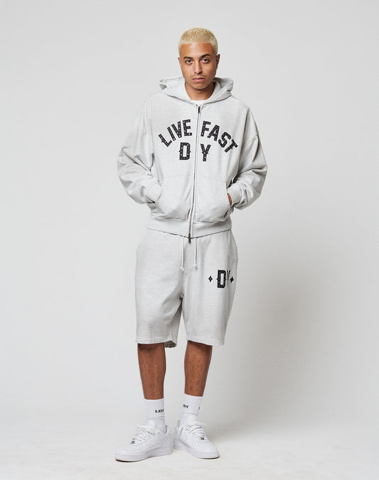 A person with short blond hair wears a light gray LIVE FAST DIE YOUNG hoodie with hand-crafted spray effect, DY Rivet Shorts featuring a small logo, white socks, and white sneakers, standing against a plain white background.
