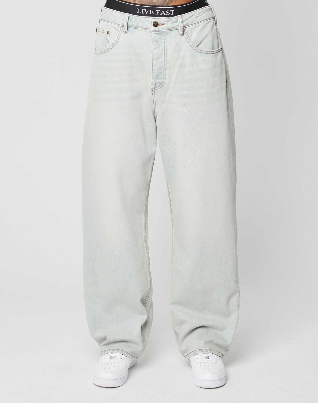 A person wears LFDY Baggy Denim by LIVE FAST DIE YOUNG in a baggy fit light-wash style, paired with white sneakers and a black LIVE FAST waistband, standing against a plain white background.