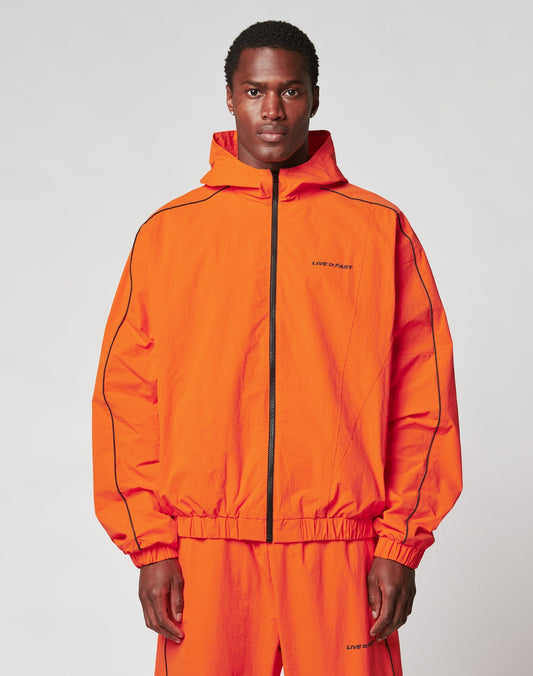A man faces forward against a plain background, wearing the LIVE FAST DIE YOUNG Live Fast Trackjacket and matching pants in bright orange Nylon-Elastan blend, with elastic cuffs, black piping, and small text on the left chest and leg.