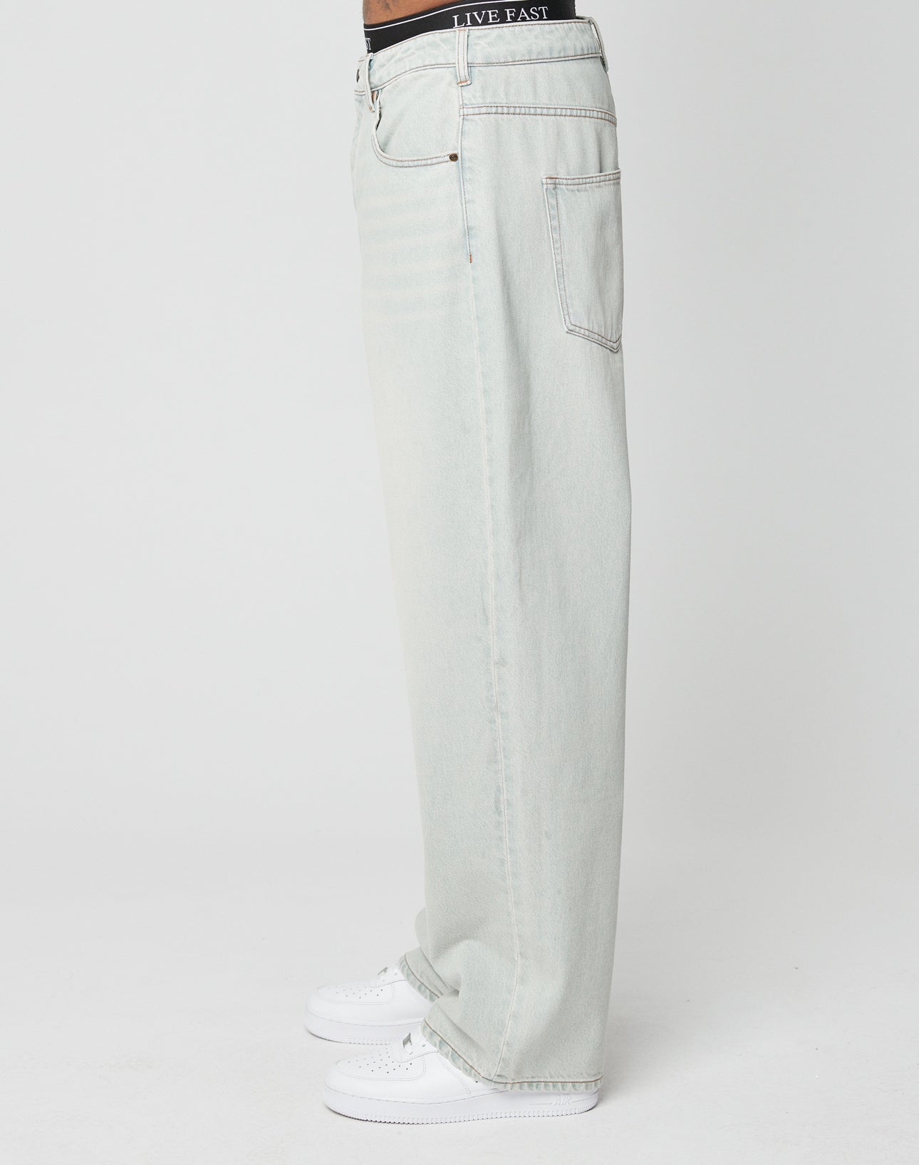 A person in profile wears LIVE FAST DIE YOUNG’s LFDY Baggy Denim jeans in light-wash Baumwolle, paired with white sneakers. A black LIVE FAST waistband shows above the jeans. The background is plain white.