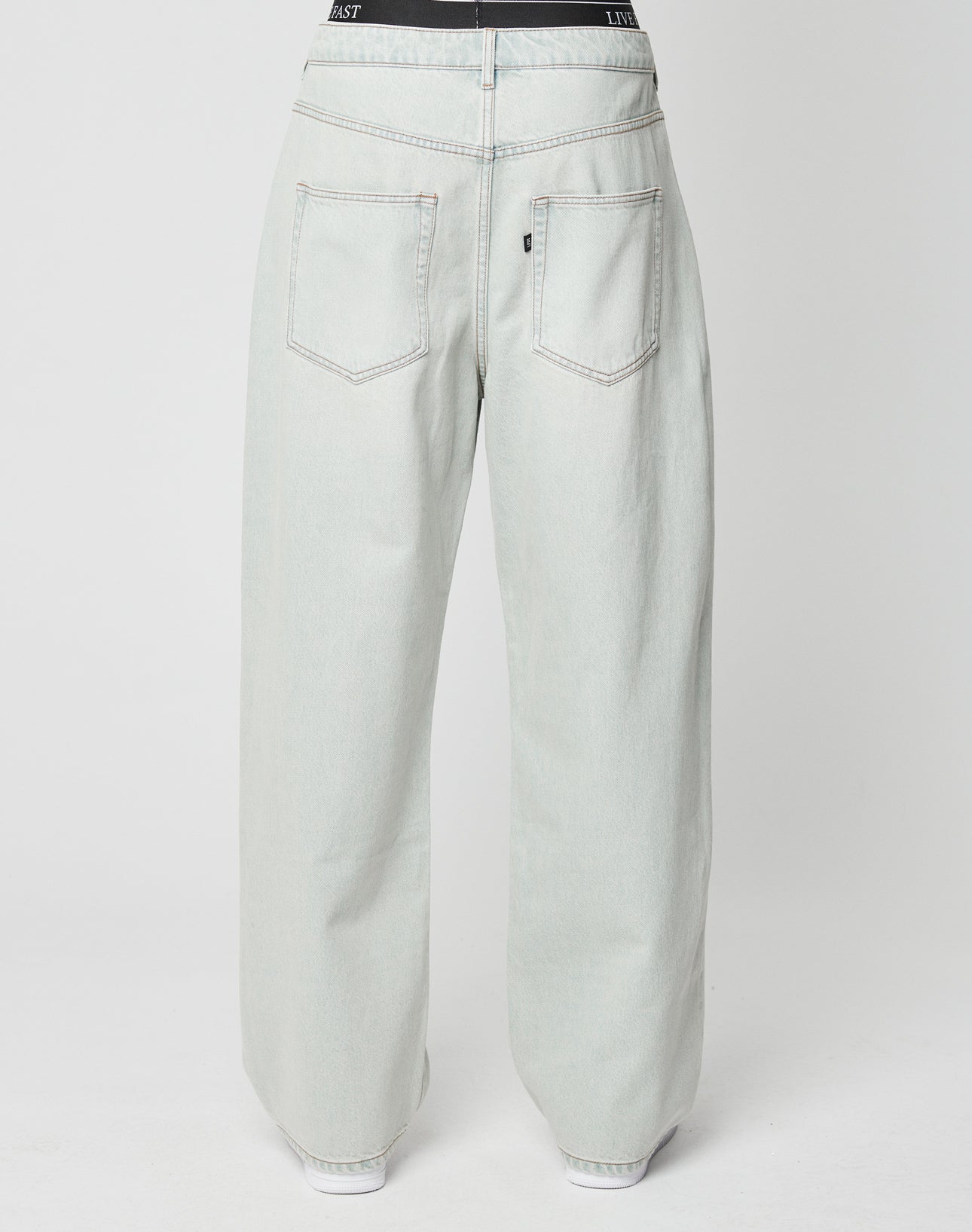 Back view of a person wearing LFDY Baggy Denim by LIVE FAST DIE YOUNG—high-waisted, light-wash, wide-leg jeans in a baggy fit with two large back pockets and a black waistband. White sneakers finish the look against a plain white background.