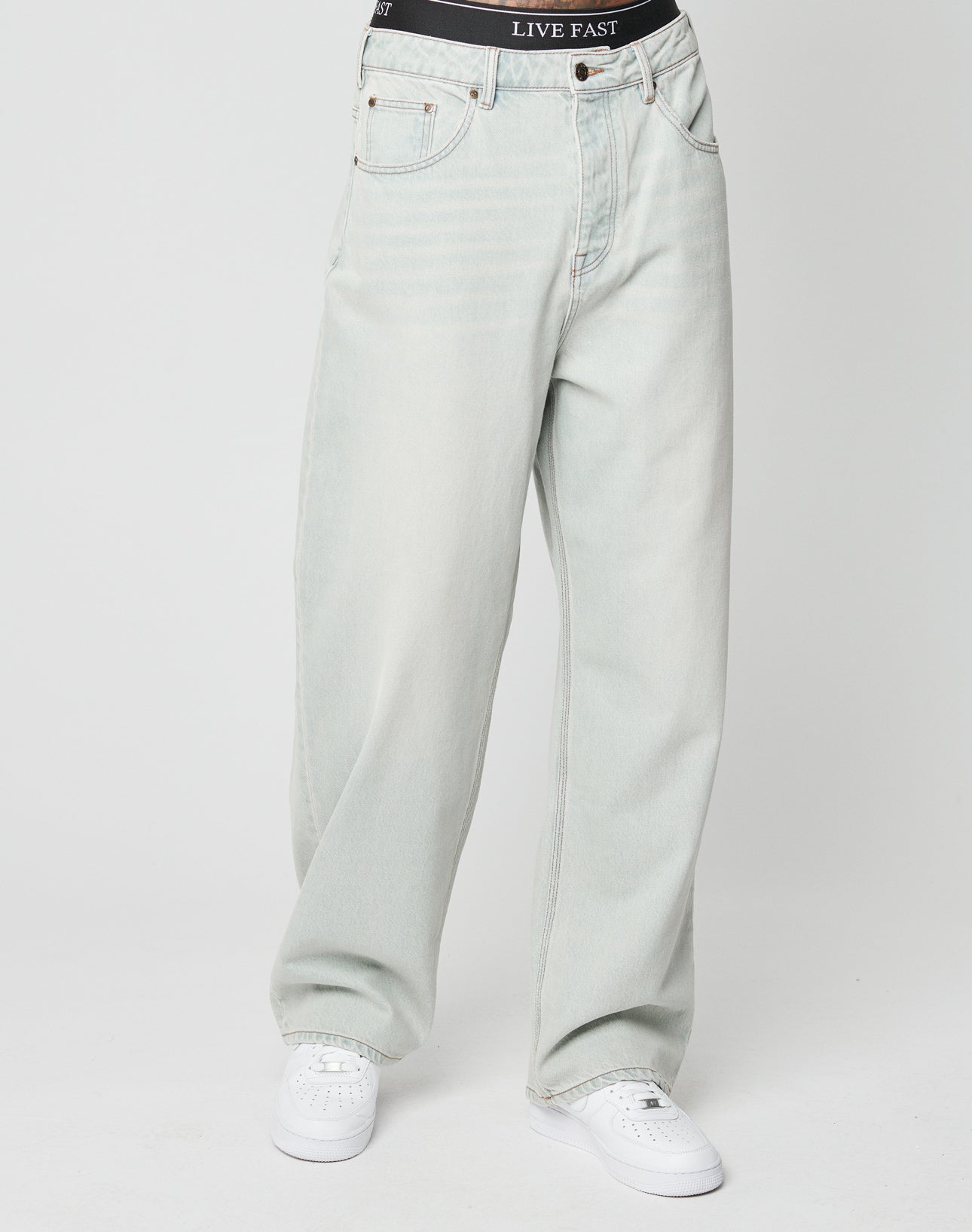 Model wears LFDY Baggy Denim by LIVE FAST DIE YOUNG—baggy fit, light-wash Baumwolle jeans with a black LIVE FAST waistband—styled simply with white sneakers against a plain white background.