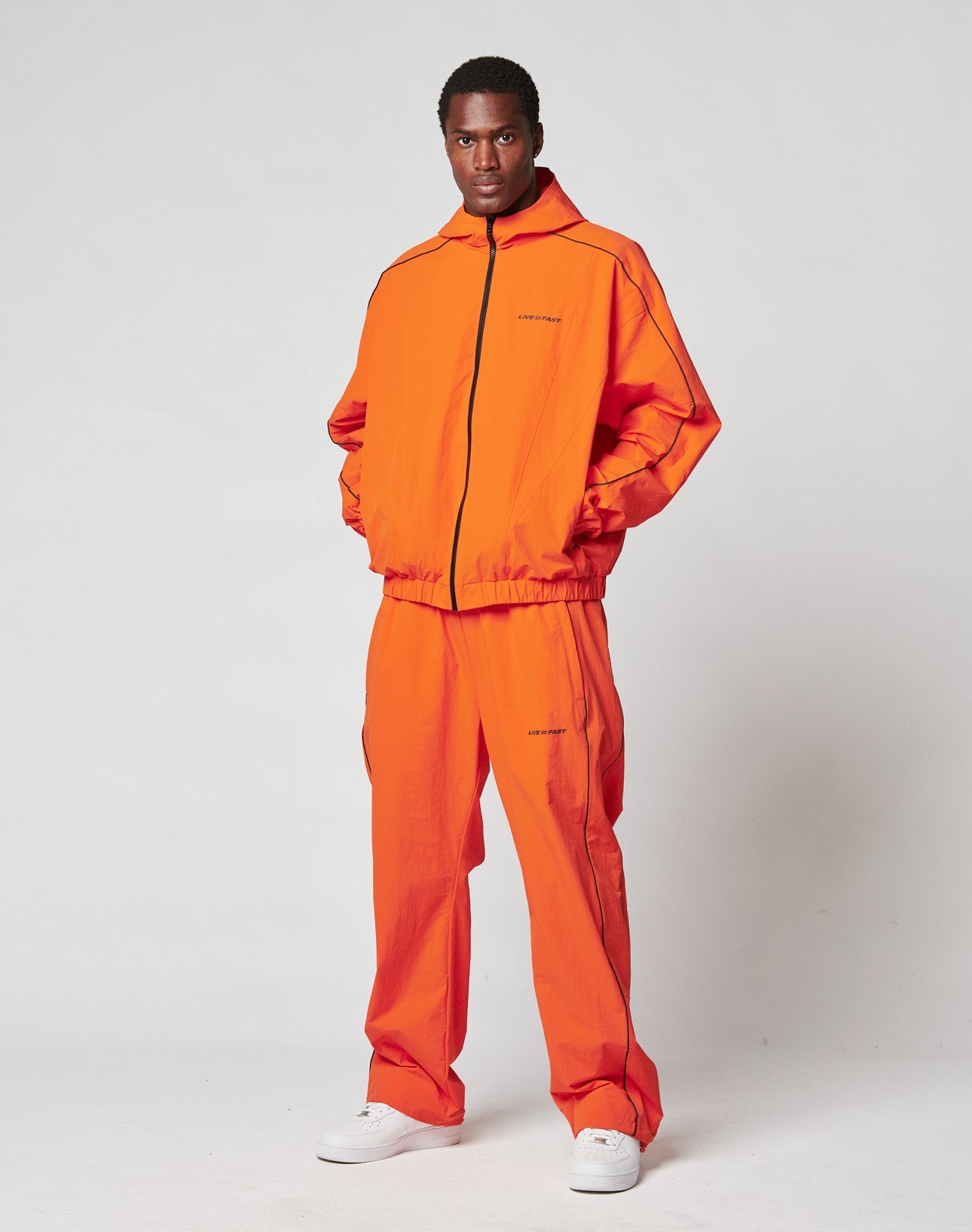 A man stands against a plain background wearing LIVE FAST DIE YOUNGs bright orange Live Fast Trackpants with black piping, reflektierender Logo Print, and Breiter Elastik-Bund. He pairs them with white sneakers and looks directly at the camera, hands in pockets.
