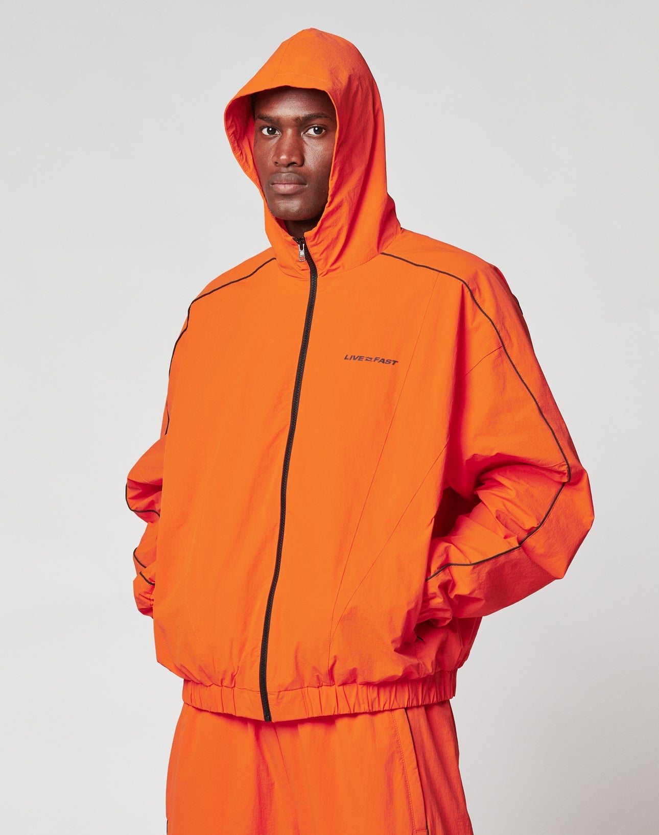 A man stands with his hands in his pockets, wearing the LIVE FAST DIE YOUNG Live Fast Trackjacket in orange with black piping. The jacket has a small LIVE FAST logo, elastische Bündchen, and is made from a durable Nylon-Elastan Mischung.