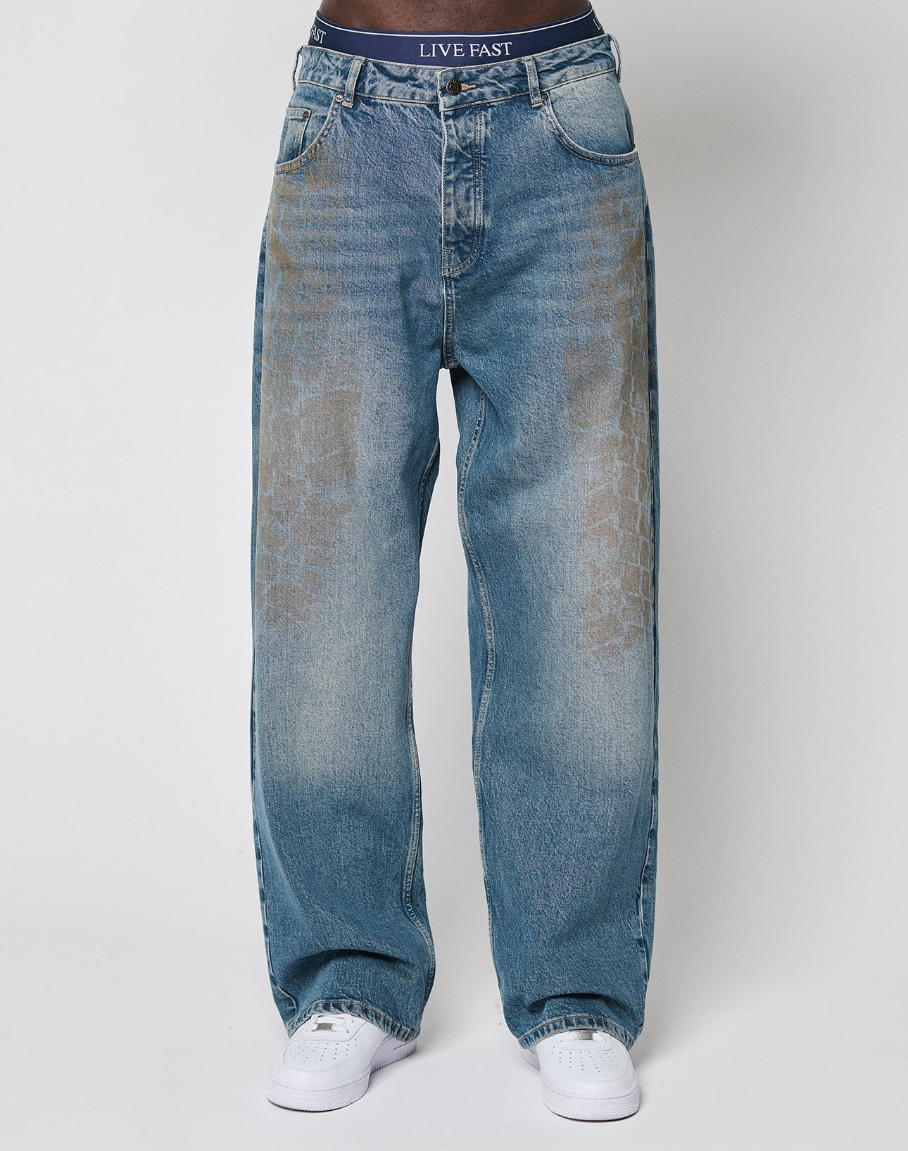 A person wears LFDY Tire Tracks Denim by LIVE FAST DIE YOUNG, featuring a baggy fit in faded blue with a subtle tire track pattern and visible branded waistband above the jeans, paired with white sneakers against a plain white background.
