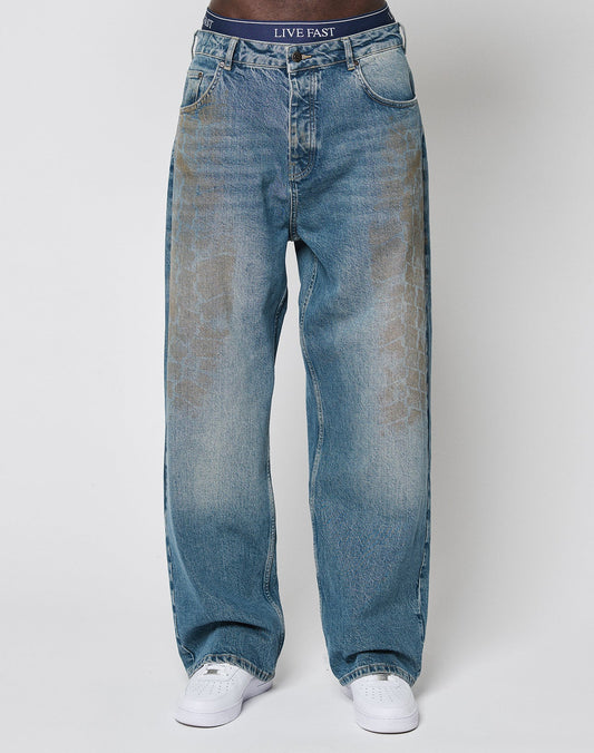 A person wears LFDY Tire Tracks Denim by LIVE FAST DIE YOUNG, featuring a baggy fit in faded blue with a subtle tire track pattern and visible branded waistband above the jeans, paired with white sneakers against a plain white background.
