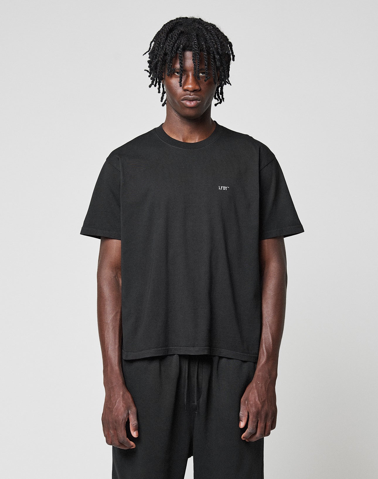 A person with short, twisted hair faces forward wearing the LIVE FAST DIE YOUNG Basic Chest Tee in black and black pants, standing against a neutral light gray background. | model