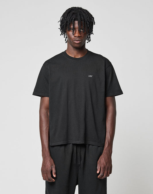A person with short, twisted hair faces forward wearing the LIVE FAST DIE YOUNG Basic Chest Tee in black and black pants, standing against a neutral light gray background. | model