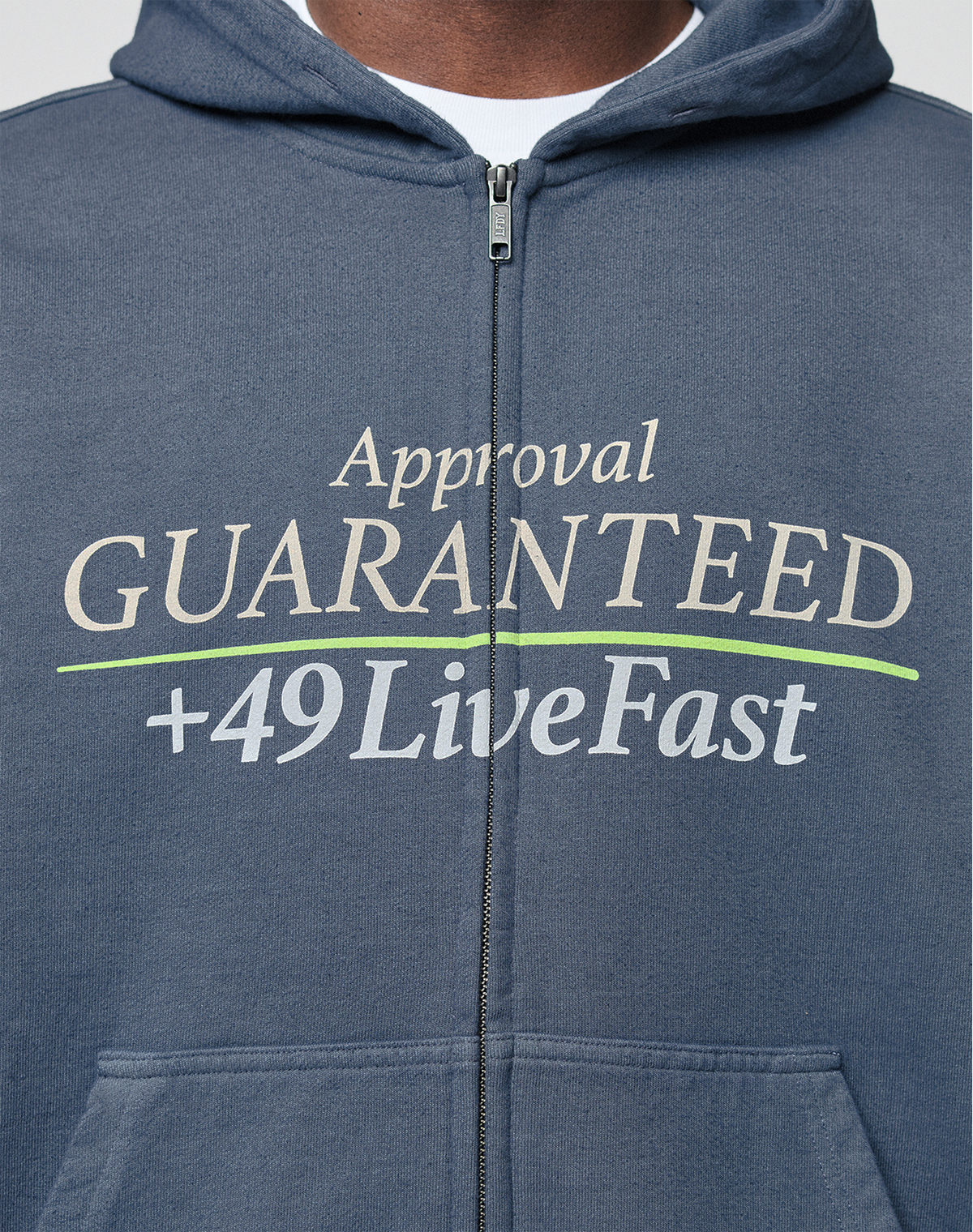 Close-up of the LIVE FAST DIE YOUNG LFDY Guaranteed Hooded Zip in blue, featuring frontprint white and green "Approval GUARANTEED +49 LiveFast" text. Made from 100% Baumwolle, modeled showing only chest and neck.
