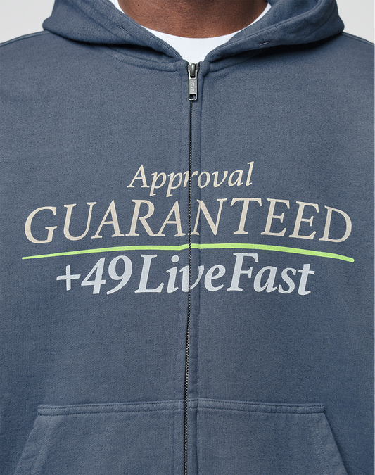 Close-up of the LIVE FAST DIE YOUNG LFDY Guaranteed Hooded Zip in blue, featuring frontprint white and green "Approval GUARANTEED +49 LiveFast" text. Made from 100% Baumwolle, modeled showing only chest and neck.