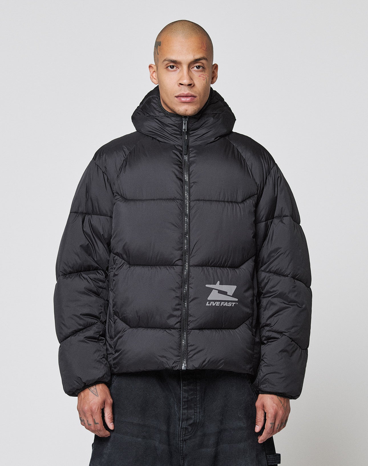 A person with a shaved head wears the LIVE FAST DIE YOUNG Live Fast Puffer Jacket in Oversize Fit, featuring a hood, a small graphic, and “LIVE FAST” lettering on the left waist, set against a plain light background.