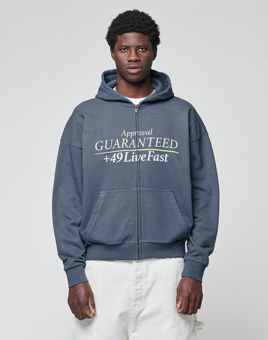 A man stands against a light gray background wearing the LFDY Guaranteed Hooded Zip by LIVE FAST DIE YOUNG, featuring an oversized dark blue fit, frontprint "Approval GUARANTEED +49 Live Fast", 100% Baumwolle. He pairs it with light-colored pants. | model