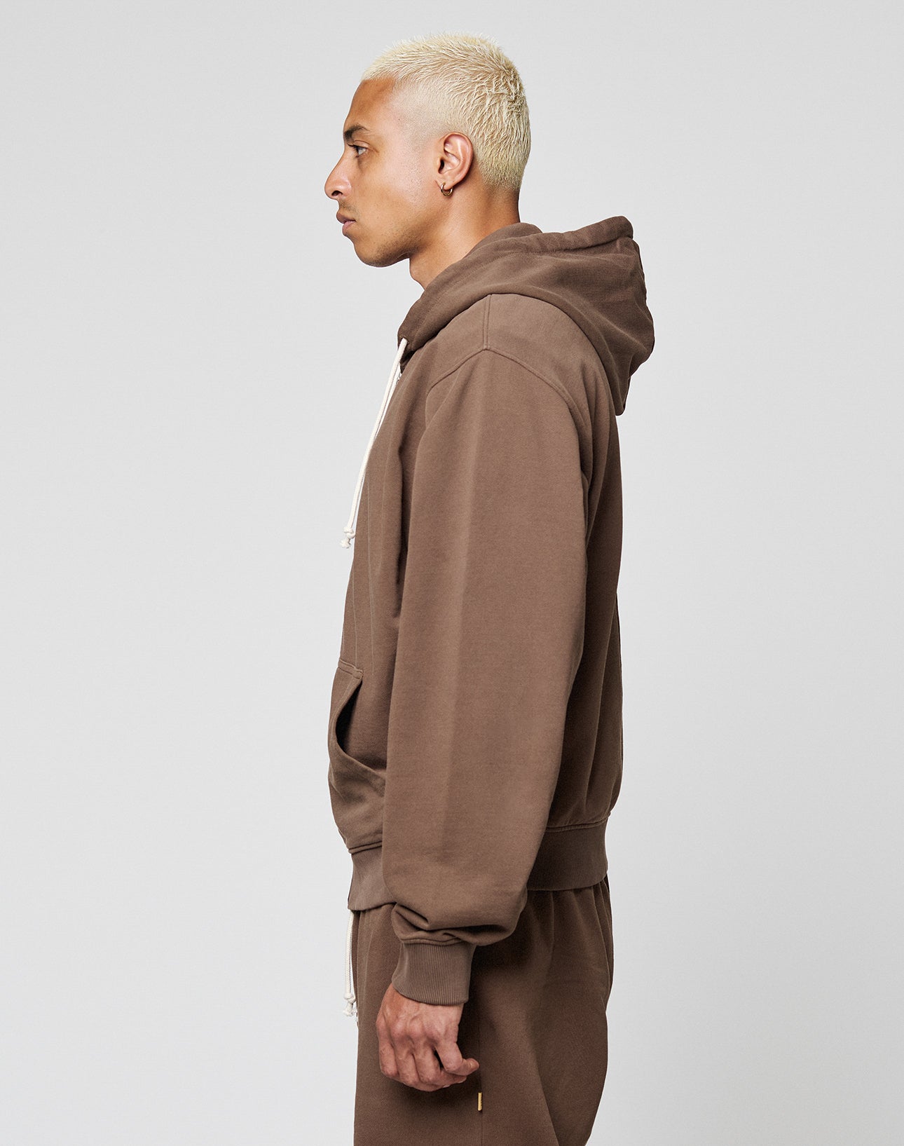 A person with short bleached blonde hair wears the LIVE FAST DIE YOUNG LFDY Lightweight Hooded Zip in brown and matching pants, Regular Fit, standing in profile against a plain light gray background.