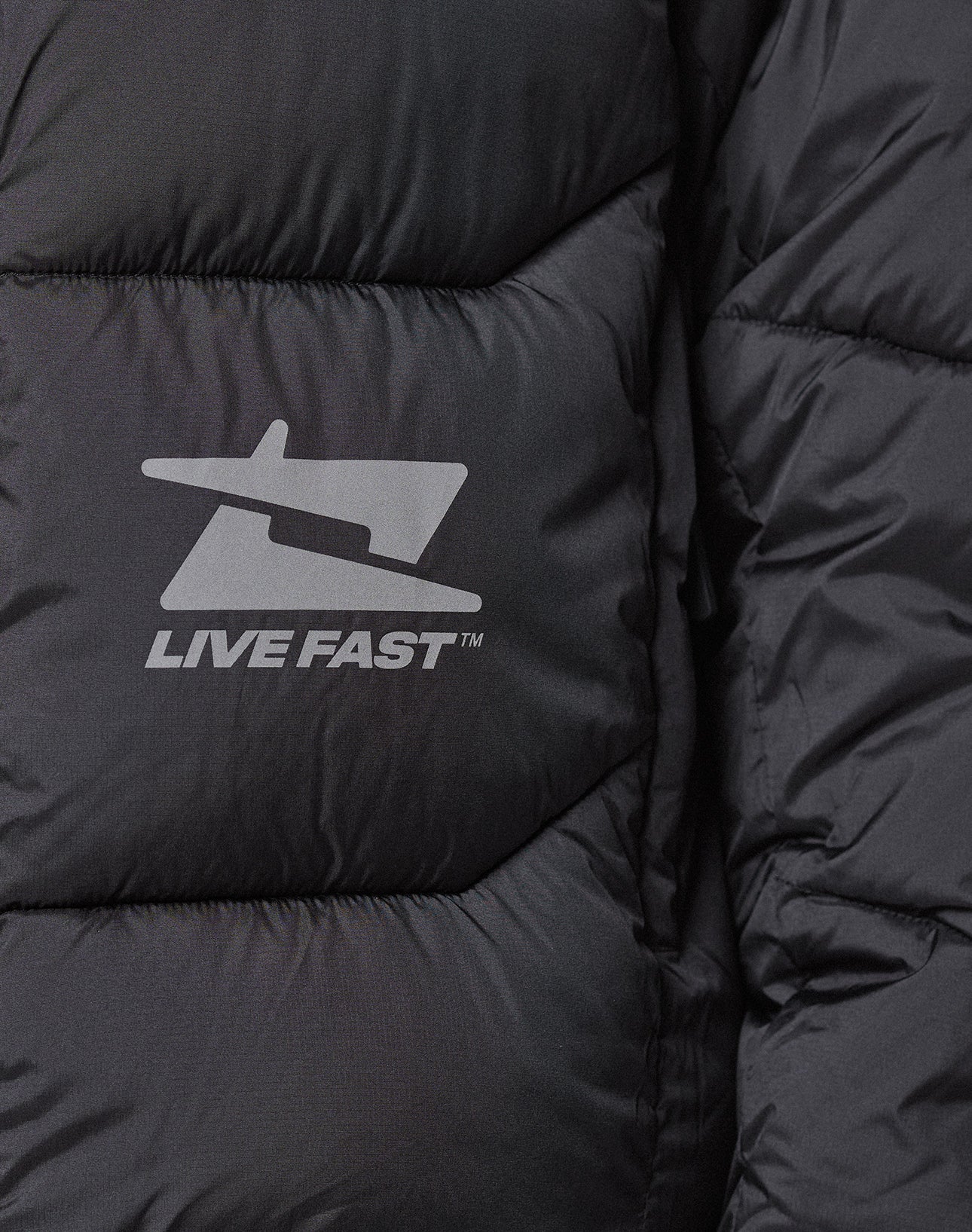 Close-up of the Live Fast Puffer Jacket by LIVE FAST DIE YOUNG, a black quilted jacket with a stylized jet logo and LIVE FAST™ in white reflektierender Print for enhanced visibility.