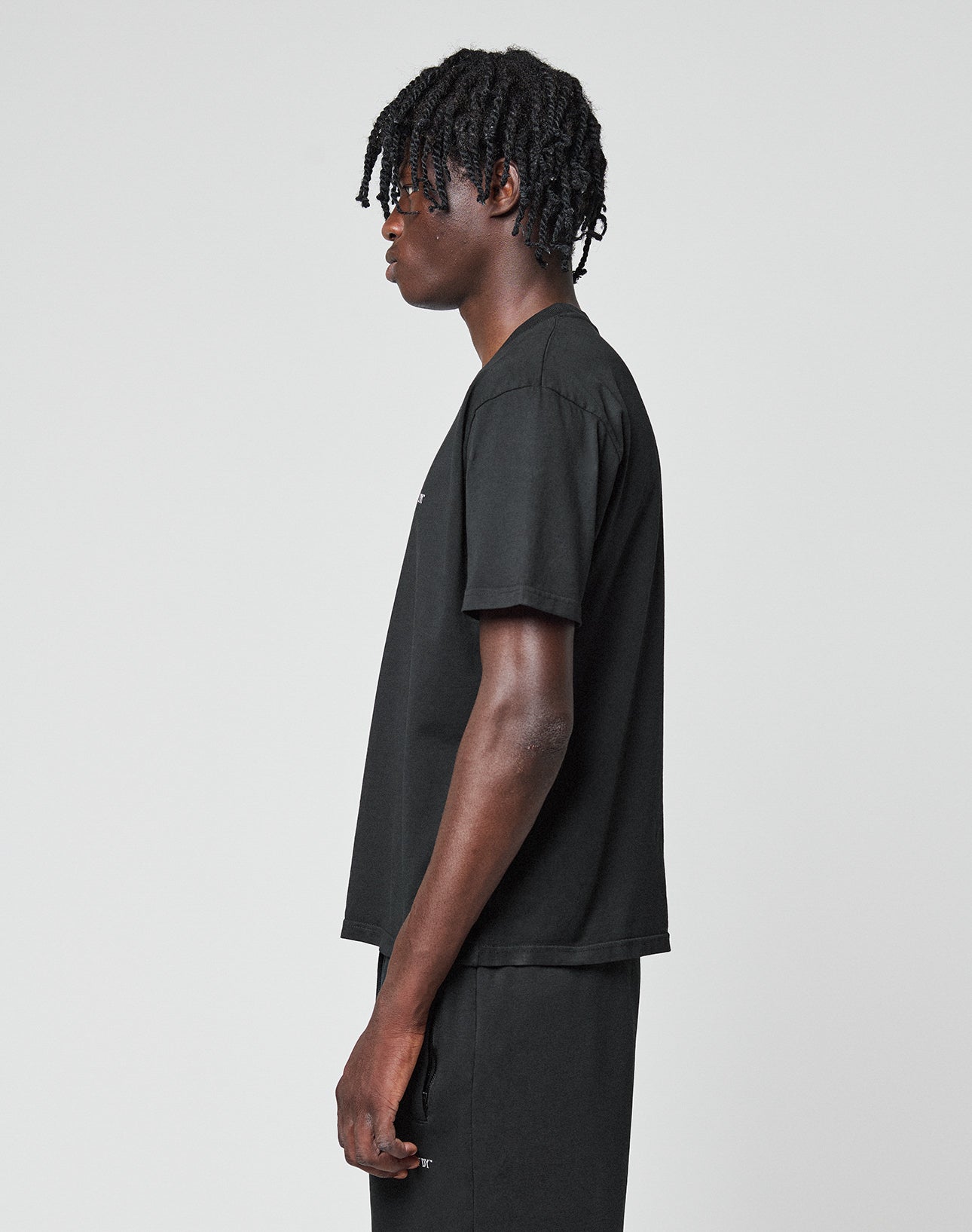A person with short, twisted hair stands in profile against a plain background, wearing the LIVE FAST DIE YOUNG Basic Chest Tee—a regular-fit black T-shirt with a subtle logo print—paired with matching black Baumwolle pants.