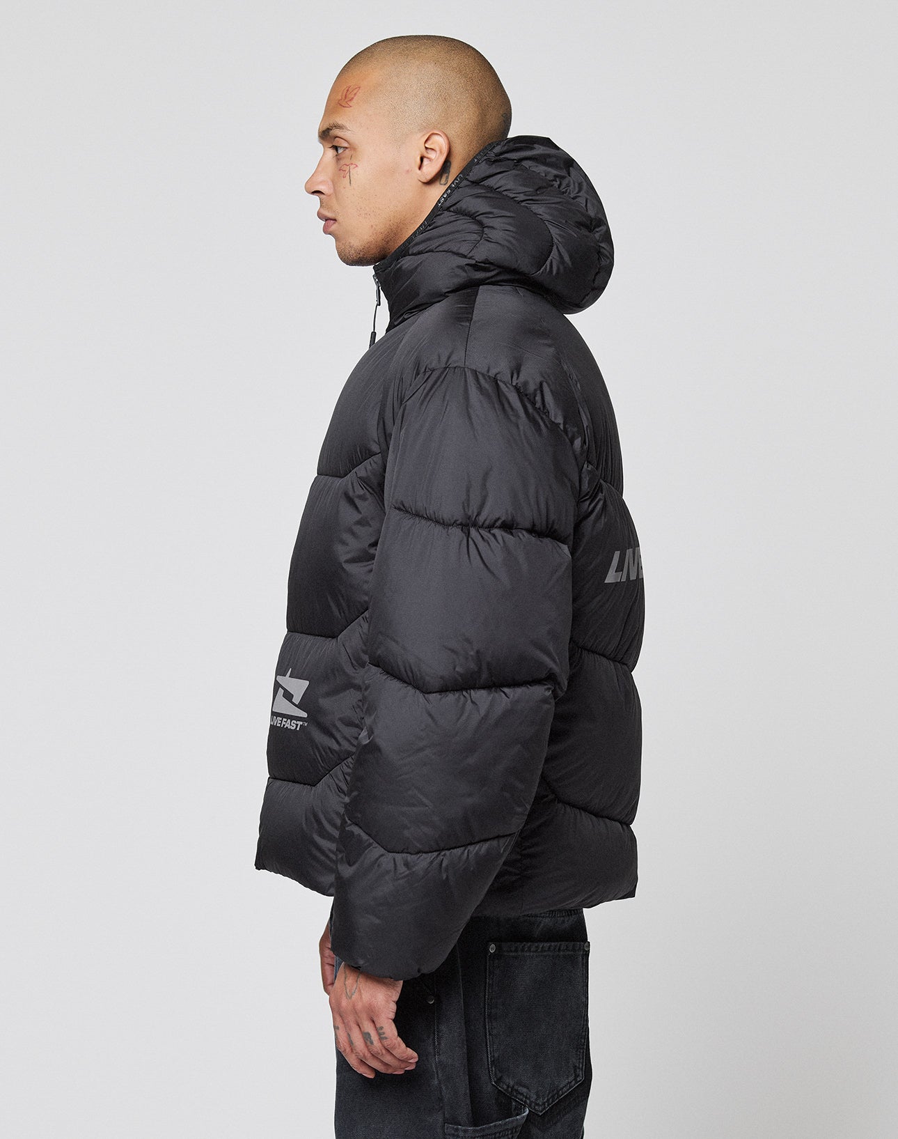 A person with a shaved head wears the LIVE FAST DIE YOUNG Live Fast Puffer Jacket in oversize fit black with a hood, featuring a small logo on the left sleeve and a reflective print on the side, standing in profile against a white background.