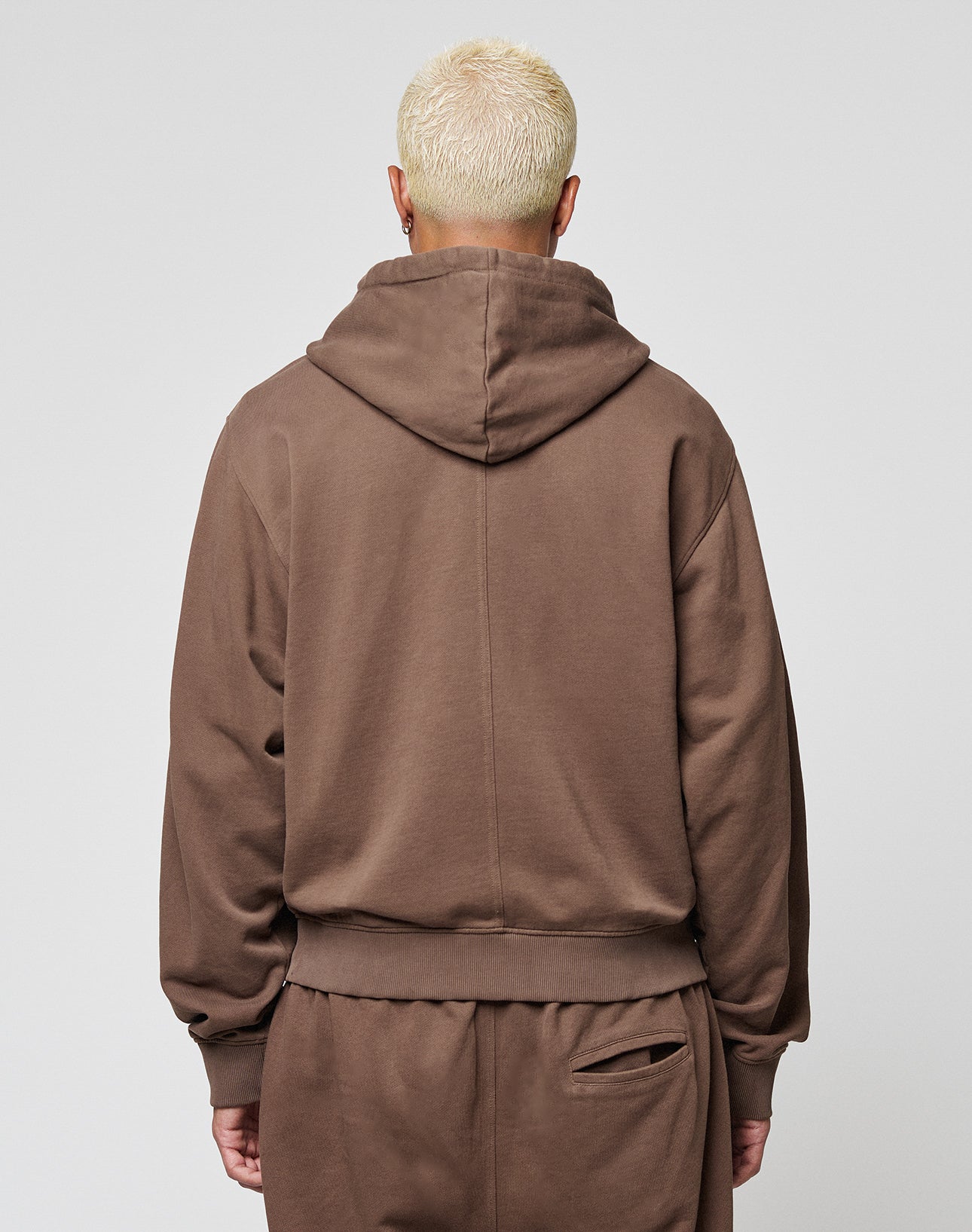 Seen from behind, a person with short, bleached blonde hair wears the LFDY Lightweight Hooded Zip by LIVE FAST DIE YOUNG and matching brown Regular Fit pants, standing against a plain light gray background.