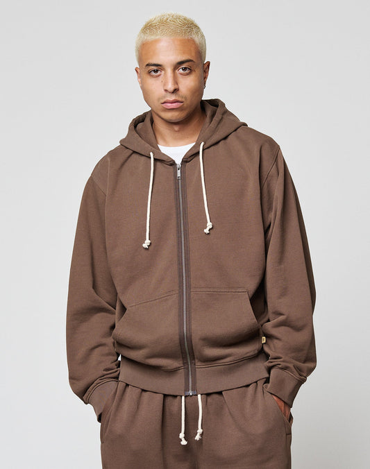 A person with short, bleached blond hair wears the LIVE FAST DIE YOUNG LFDY Lightweight Hooded Zip in brown with cream drawstrings and matching pants, standing against a plain light gray background.