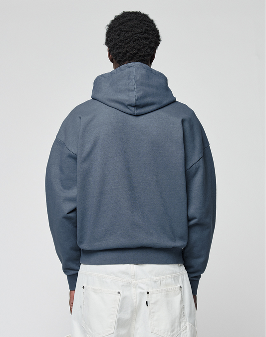 A person with short curly hair stands with their back to the camera in the LIVE FAST DIE YOUNG LFDY Guaranteed Hooded Zip, an oversize-fit blue hoodie made from 100% Baumwolle, styled with white pants featuring visible back pockets against a plain light background.