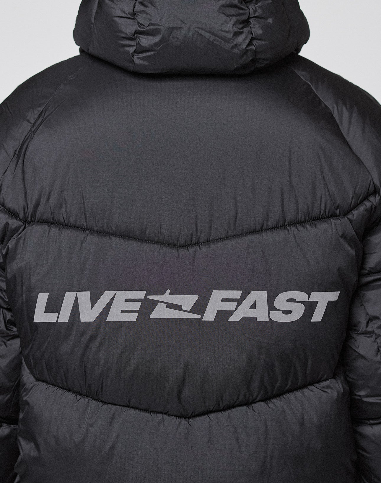 A close-up of the Live Fast Puffer Jacket by LIVE FAST DIE YOUNG, showing bold, silvery, reflective "LIVE FAST" text across the back shoulders.