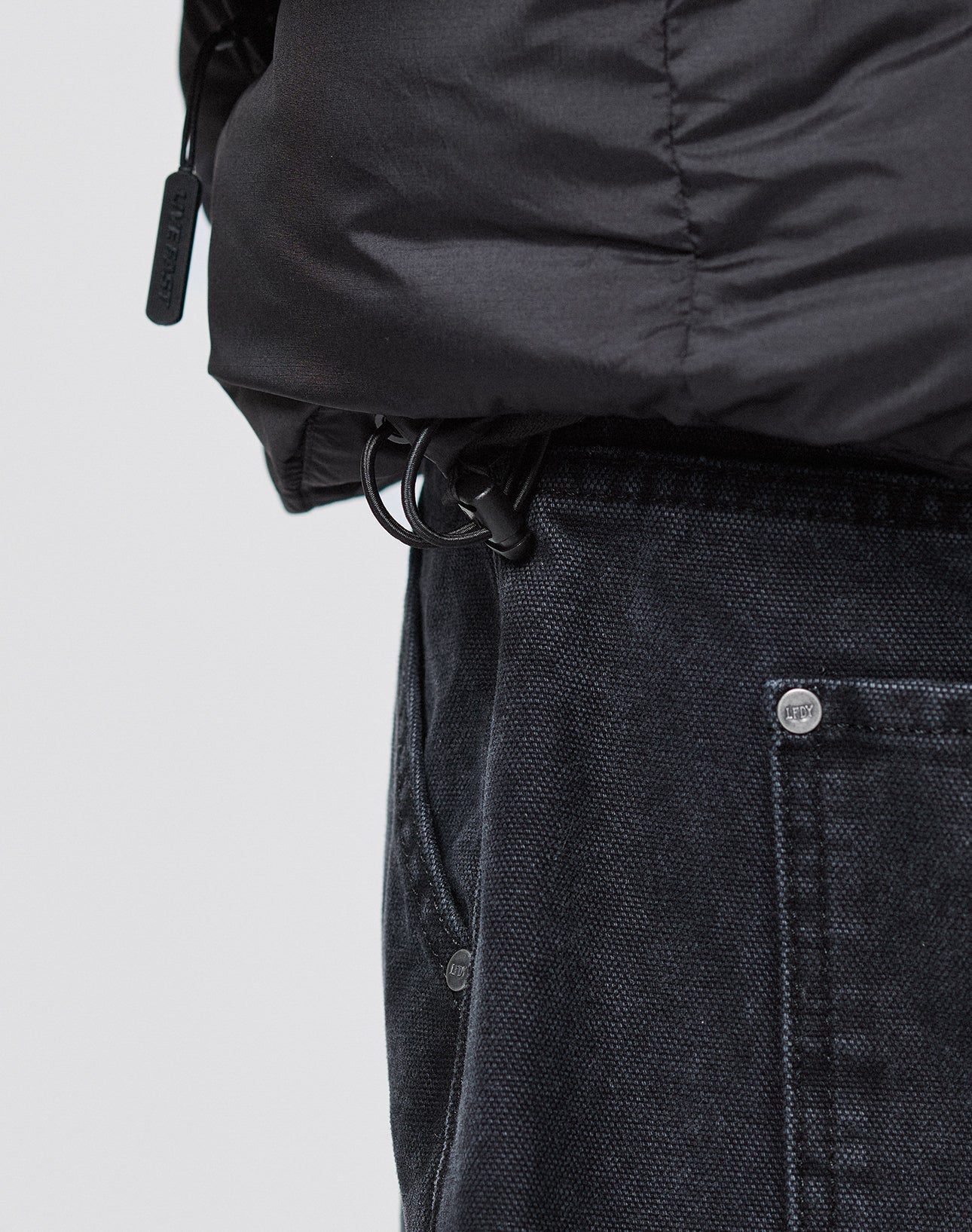Close-up of a person wearing dark denim jeans and the LIVE FAST DIE YOUNG Live Fast Puffer Jacket in an oversized fit, featuring a drawstring with plastic toggle near the waistband and large visible front pocket.