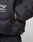 Close-up of a person in the LIVE FAST DIE YOUNG Live Fast Puffer Jacket, black with an oversize fit, visible zipper pocket, and reflective FAST logo print. The hand is tucked into a side pocket, adding a modern touch.