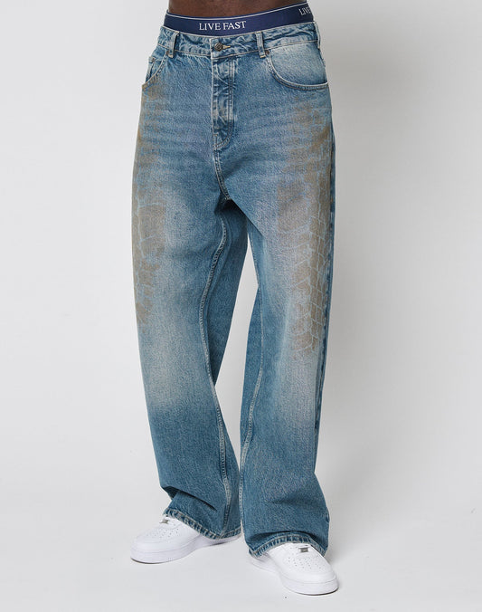 A person wearing LIVE FAST DIE YOUNG’s LFDY Tire Tracks Denim—baggy fit, light blue jeans with faded tire track patterns and a LIVE FAST waistband—paired with white sneakers, stands against a plain white background.