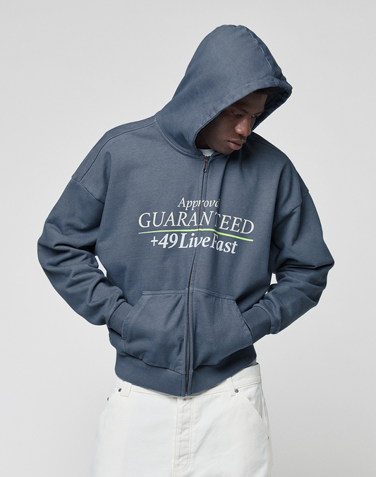 A person wears the LFDY Guaranteed Hooded Zip by LIVE FAST DIE YOUNG—an oversized blue zip-up hoodie with bold front print, 100% cotton, hood up and hands in pockets, styled with white pants while looking down.