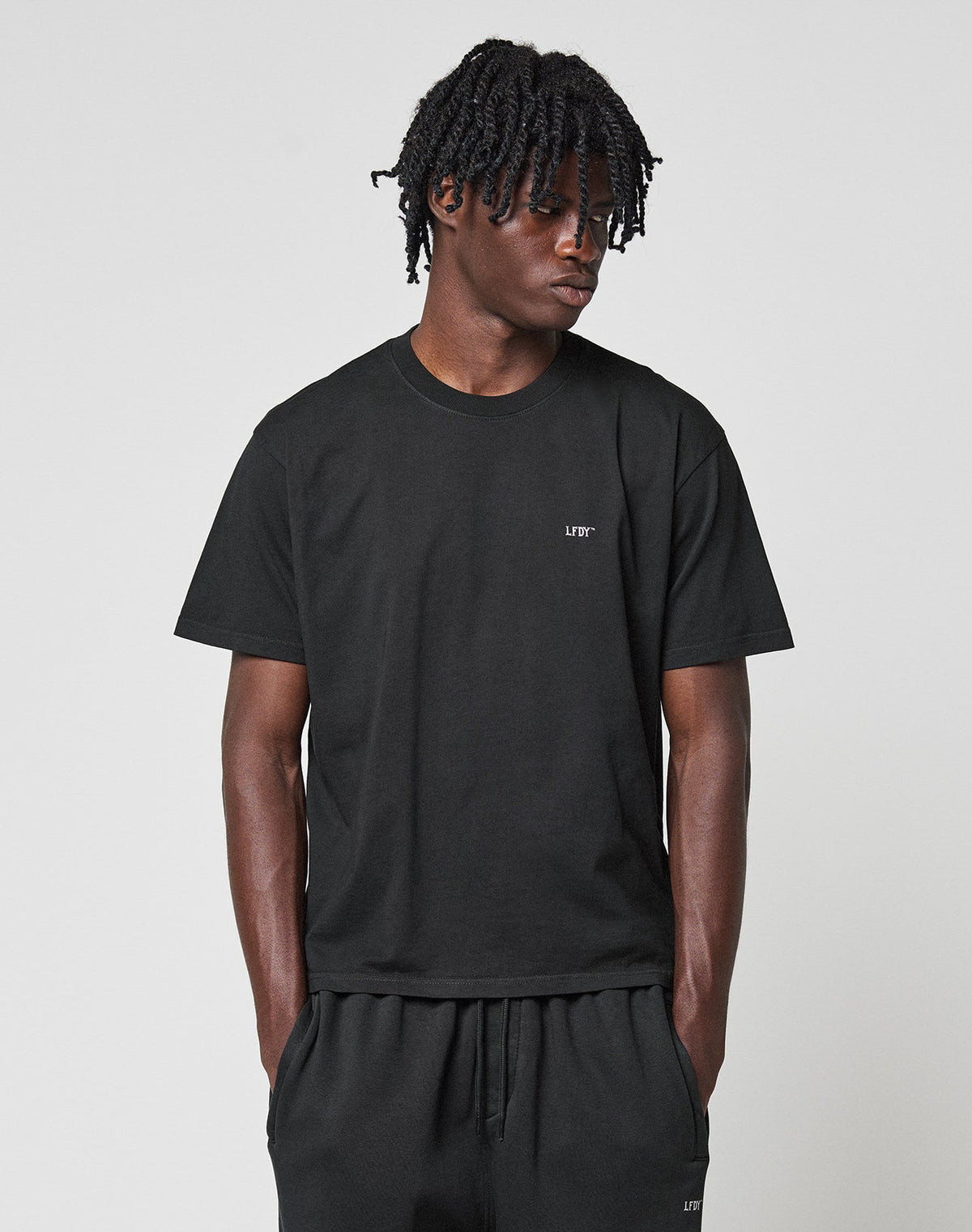 A person with short, twisted hair stands against a white background, wearing the LIVE FAST DIE YOUNG Basic Chest Tee in black and matching cotton shorts, hands in pockets and looking slightly to the side.