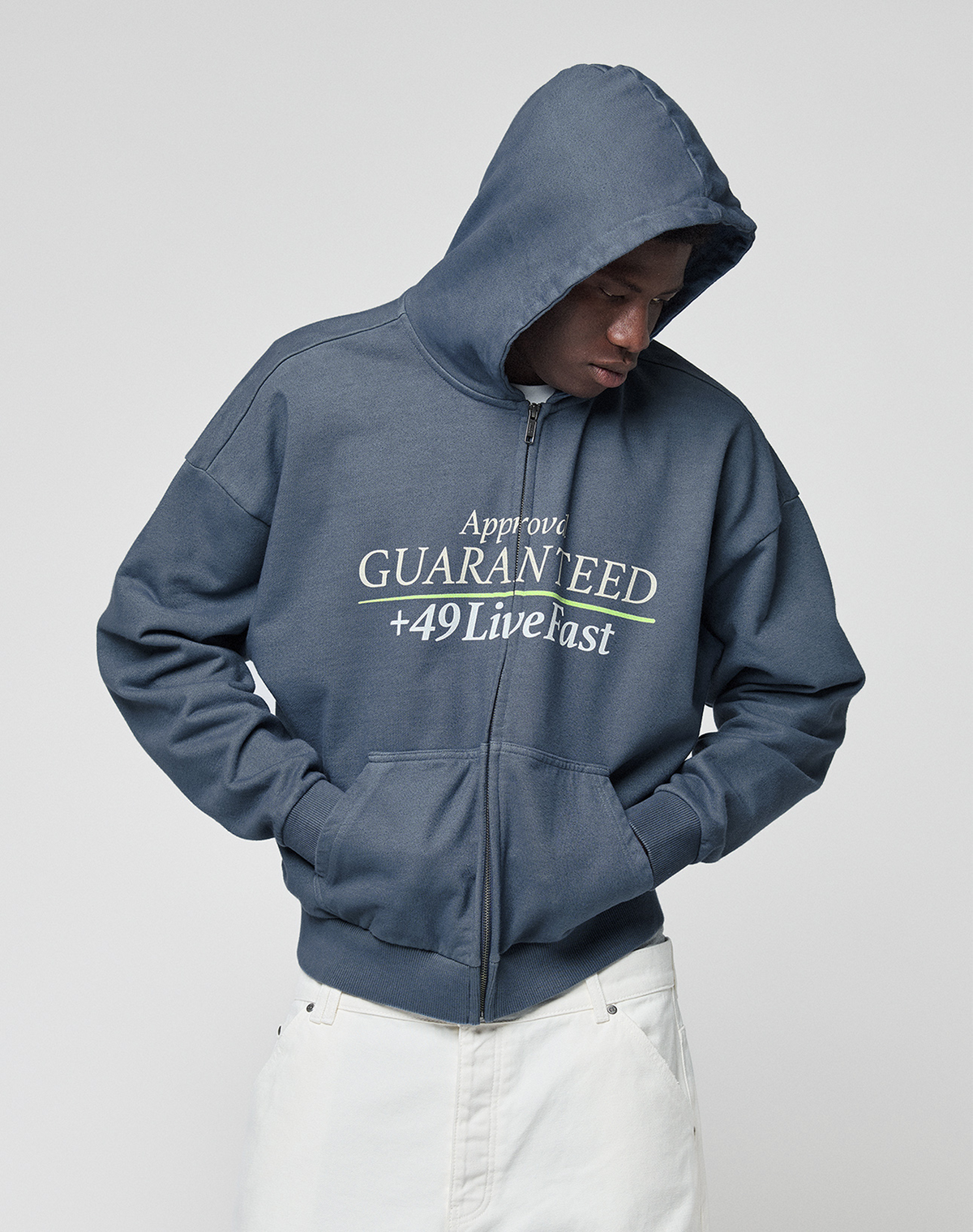 A person wears the LFDY Guaranteed Hooded Zip by LIVE FAST DIE YOUNG—an oversized blue zip-up hoodie with bold front print, 100% cotton, hood up and hands in pockets, styled with white pants while looking down.