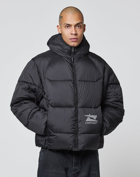 A person with a shaved head and scalp tattoos wears the LIVE FAST DIE YOUNG Live Fast Puffer Jacket in black, featuring an oversized fit, hood, front pocket arrow, LIVE FAST logo, and reflective print. Plain light gray background.