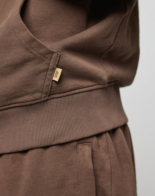 Close-up of a person in brown LFDY Lightweight Hooded Zip sweatpants by LIVE FAST DIE YOUNG, highlighting the ribbed waistband, side pocket, and a small beige tag with text on the 100% Baumwolle sweatshirt.