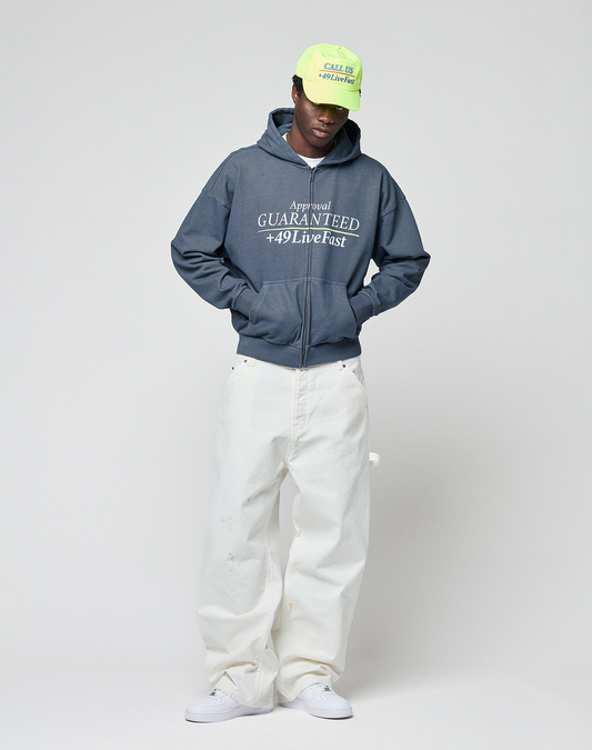 A person wearing a neon green cap, loose oversized white pants, white sneakers, and the LIVE FAST DIE YOUNG LFDY Guaranteed Hooded Zip with white frontprint stands against a plain light gray background, looking down with hands in pockets.