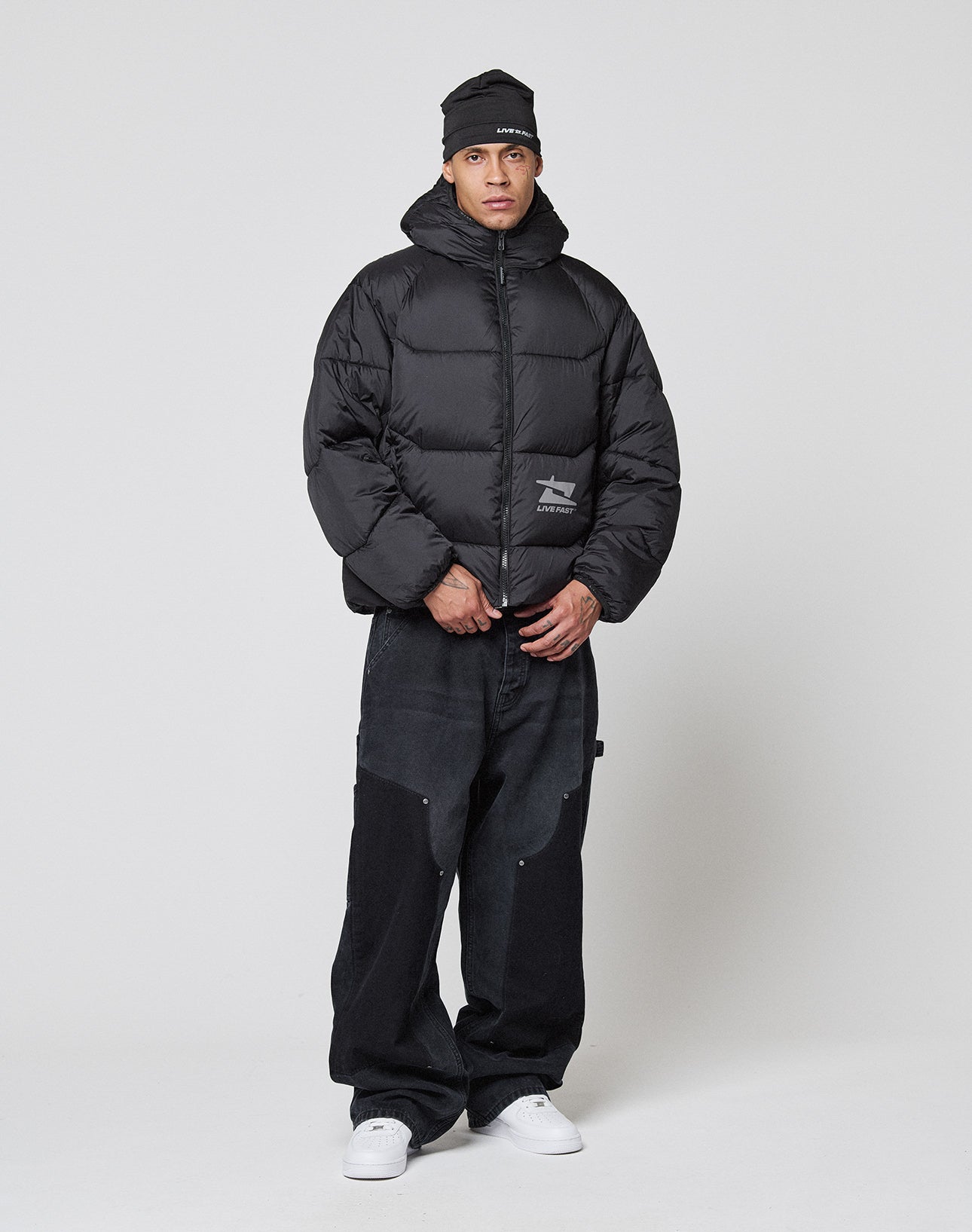 A person stands against a plain background wearing the LIVE FAST DIE YOUNG Live Fast Puffer Jacket, paired with a black beanie, loose black pants, and white sneakers for a casual, streetwear-inspired look.