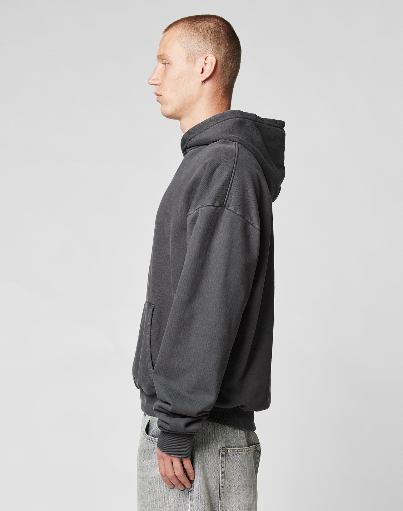 A person with short hair, wearing an Oversize Fit LFDY Core Hooded sweatshirt by LIVE FAST DIE YOUNG in dark gray and light-wash jeans, stands in profile against a plain light gray background.
