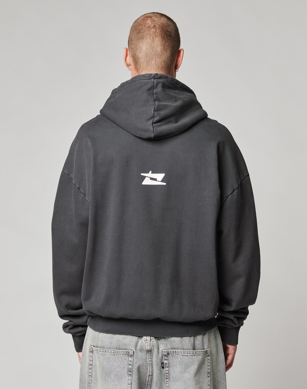 A person faces away from the camera in an oversize LFDY Core Hooded by LIVE FAST DIE YOUNG, dark cotton with a small white logo on the back. The hood is up but not worn, styled with light gray jeans.