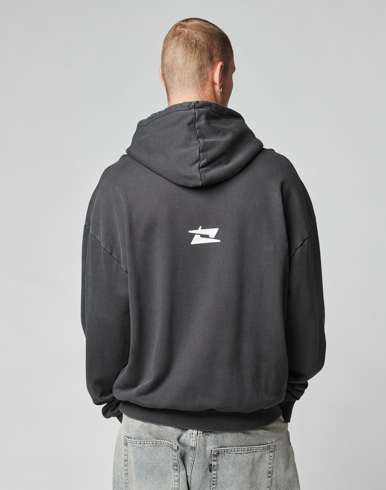A person with short hair, seen from behind, wears the LIVE FAST DIE YOUNG LFDY Core Hooded in dark cotton with a small white logo on the upper back, paired with light blue jeans against a plain gray background.