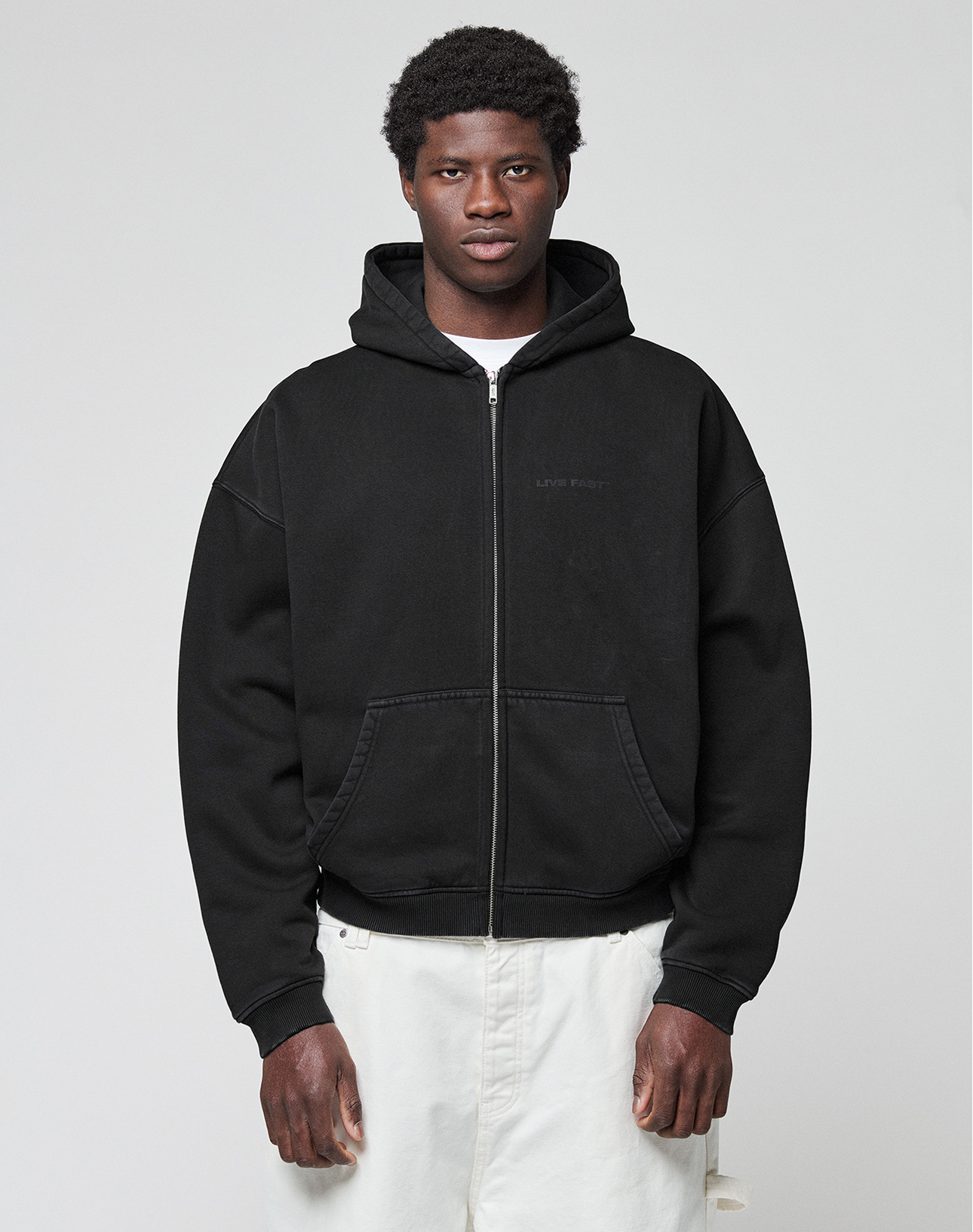 A person stands facing forward, wearing a LIVE FAST DIE YOUNG LFDY Grand Theft Hooded Zip in black and white pants, against a plain light gray background.