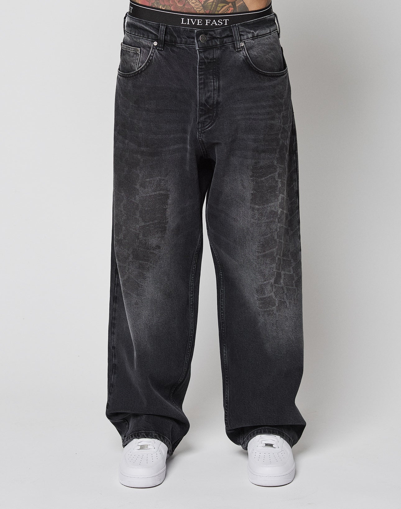 A person wears LFDY Tire Tracks Denim by LIVE FAST DIE YOUNG—loose, faded black baggy jeans with LIVE FAST on the waistband—and white sneakers, standing against a plain white background.