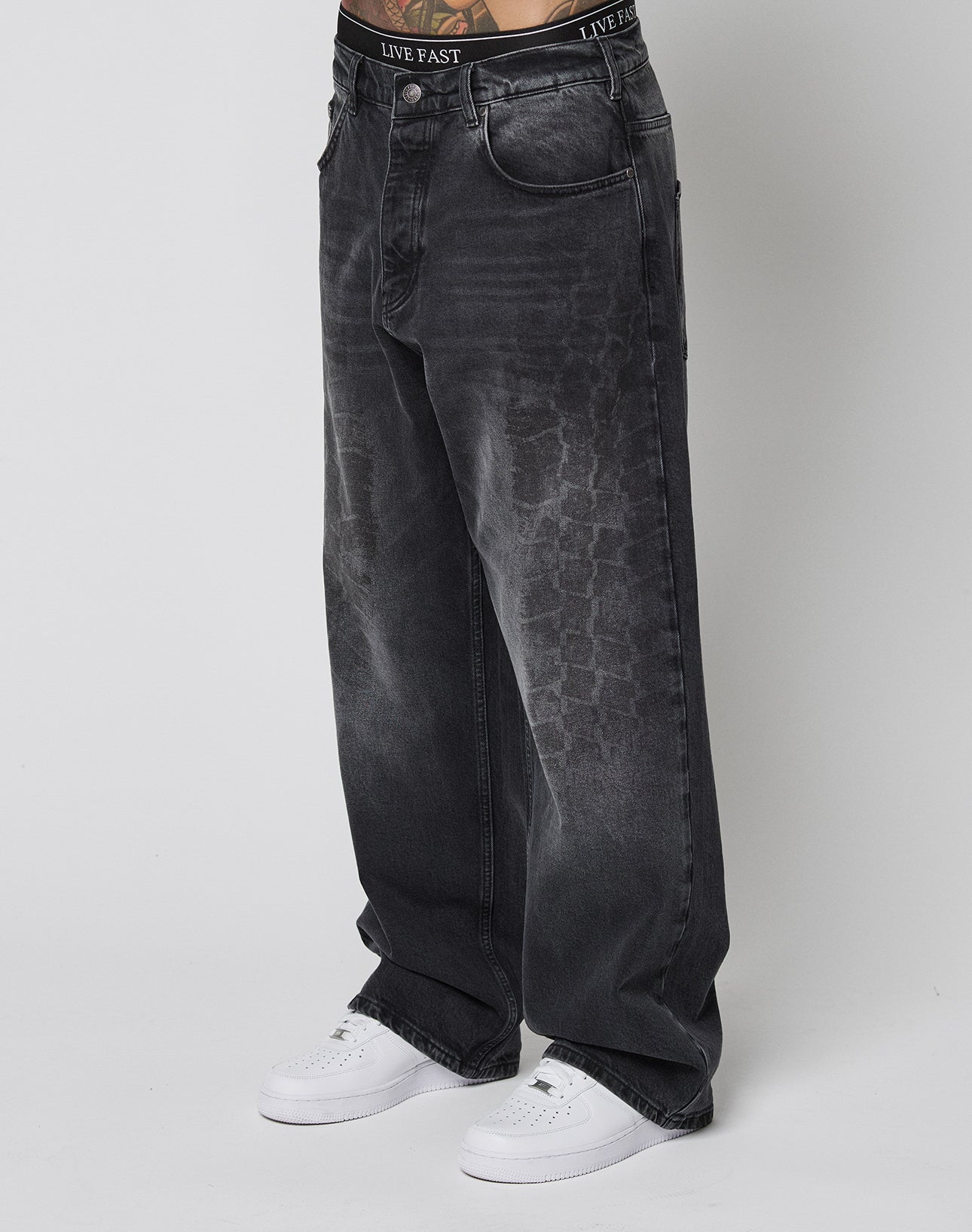 Person wearing LIVE FAST DIE YOUNG’s LFDY Tire Tracks Denim—baggy, loose-fitting, faded black jeans with a subtle tire track texture and black branded waistband—paired with white sneakers against a plain white background.
