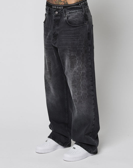 Person wearing LIVE FAST DIE YOUNG’s LFDY Tire Tracks Denim—baggy, loose-fitting, faded black jeans with a subtle tire track texture and black branded waistband—paired with white sneakers against a plain white background.