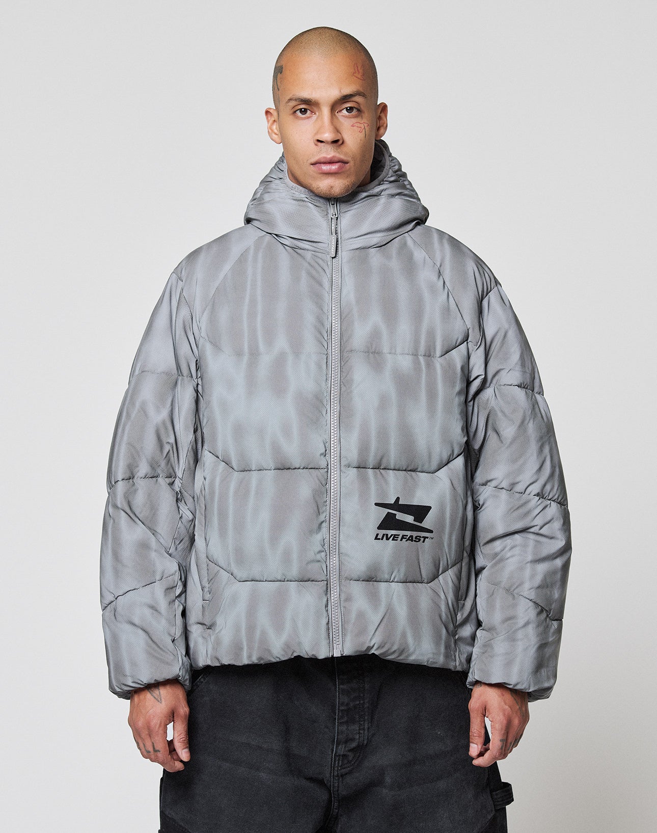 A person with a shaved head wears the LIVE FAST DIE YOUNG Live Fast Puffer Jacket, an oversized, shiny gray hooded jacket with a reflective finish and graphic logo on the lower left. The background is plain and light-colored.| model