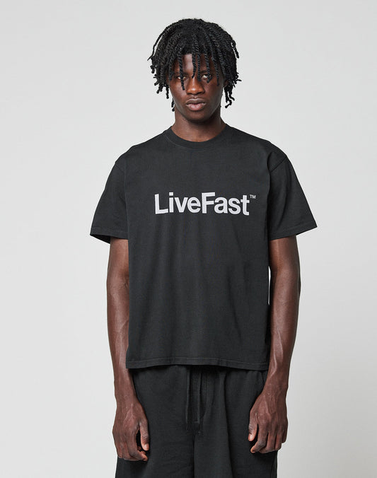 A person with short dreadlocks wears the LIVE FAST DIE YOUNG Basic LiveFast Tee in black, showcasing a bold white front print, standing against a plain light gray background. | model